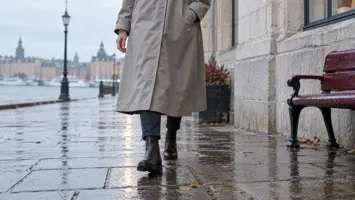 Lang umerket regnkåpe dekker lårene til en kvinne på en våt fortauskant.